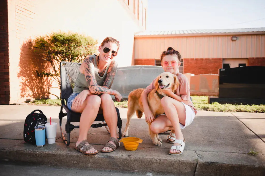 Two festival-goers with sunglasses sit outdoors with a golden retriever dog at the Dancing Rabbit Music Festival venue.