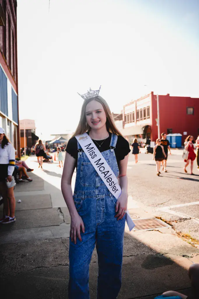 A young woman wearing a tiara and pageant sash smiles at the camera in a festival venue courtyard.