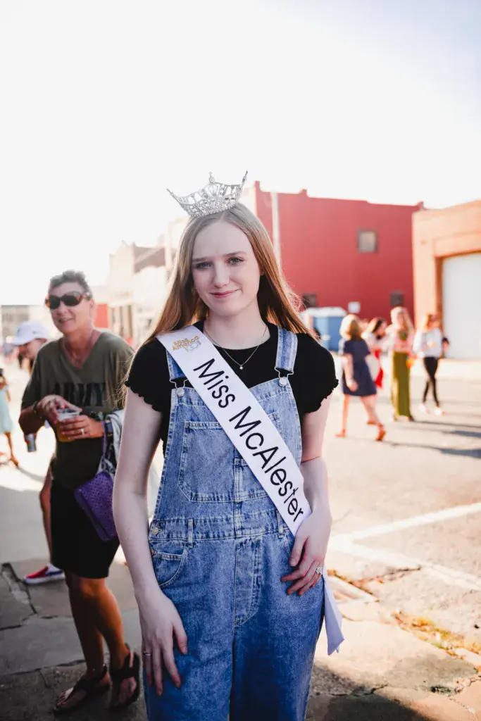 A young woman wearing a tiara and sash stands at a festival venue with colorful buildings and crowd in the background.