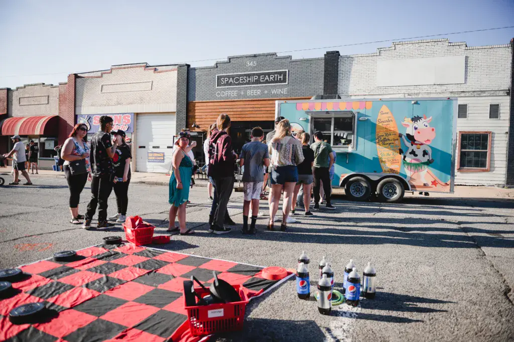 Festival attendees gather around a colorful food truck and outdoor games in a festival venue area.