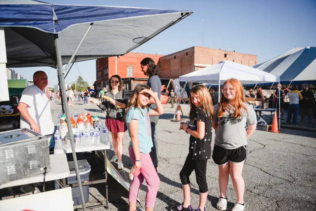 Festival attendees gather at a vendor tent with refreshments under shade structures on a sunny day.