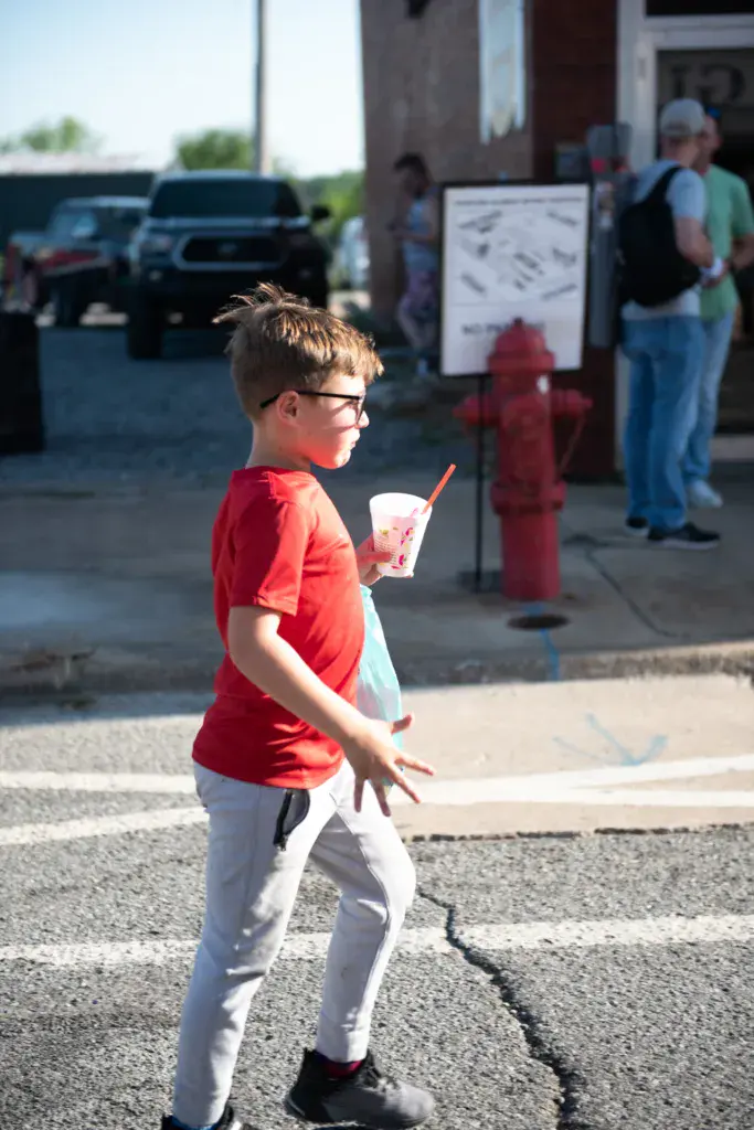 A young child in a red shirt enjoys a beverage while standing in a festival venue parking area.