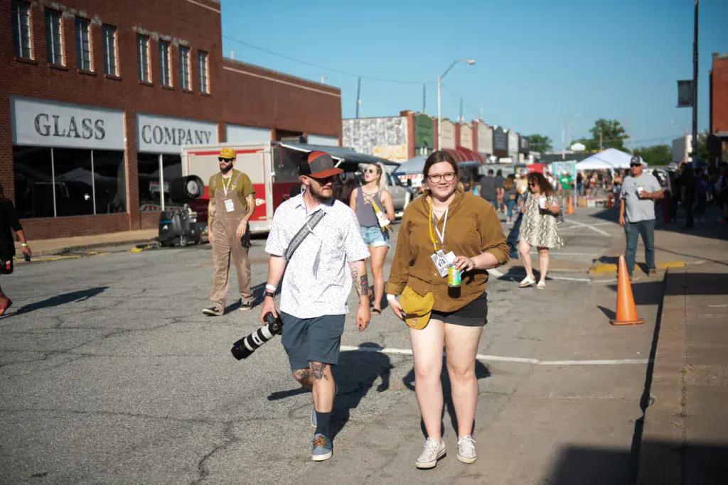Festival attendees walk through a downtown parking lot venue with brick buildings and vendor tents visible in the background.