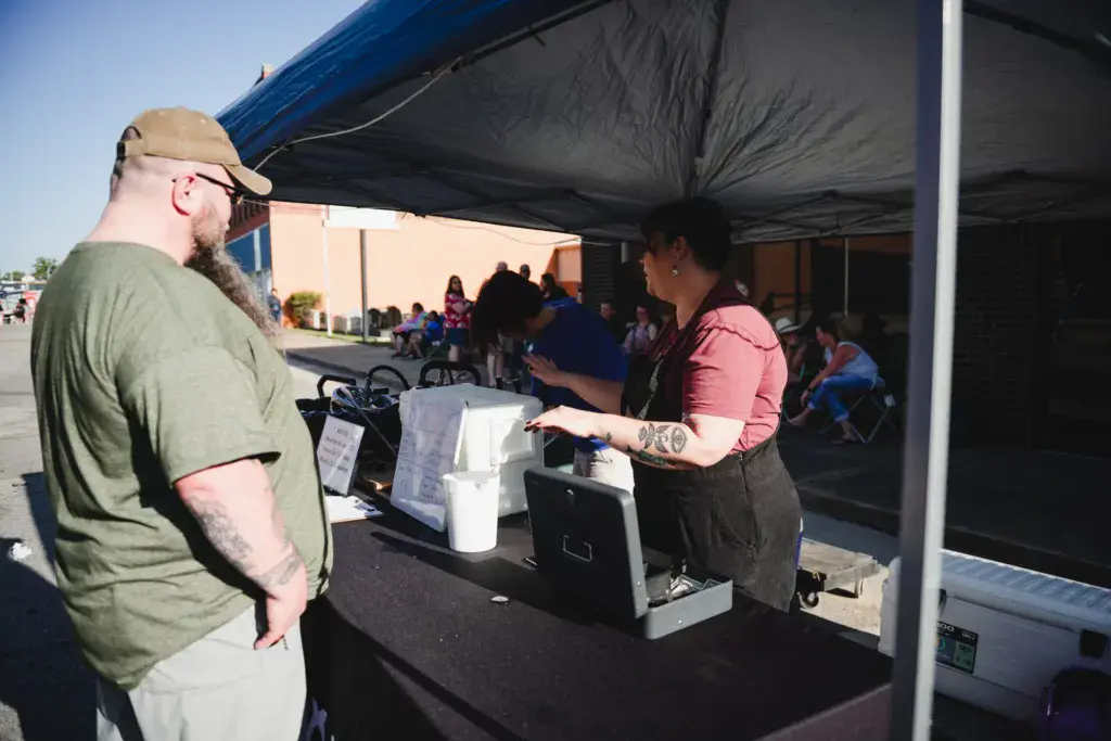 Festival vendor serves customers at an outdoor tent booth during daylight hours.