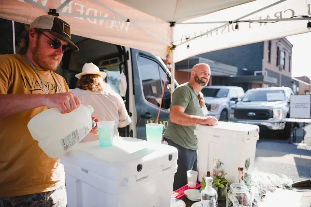 Two men serve drinks under a tent canopy at an outdoor festival venue with vehicles visible in the background.