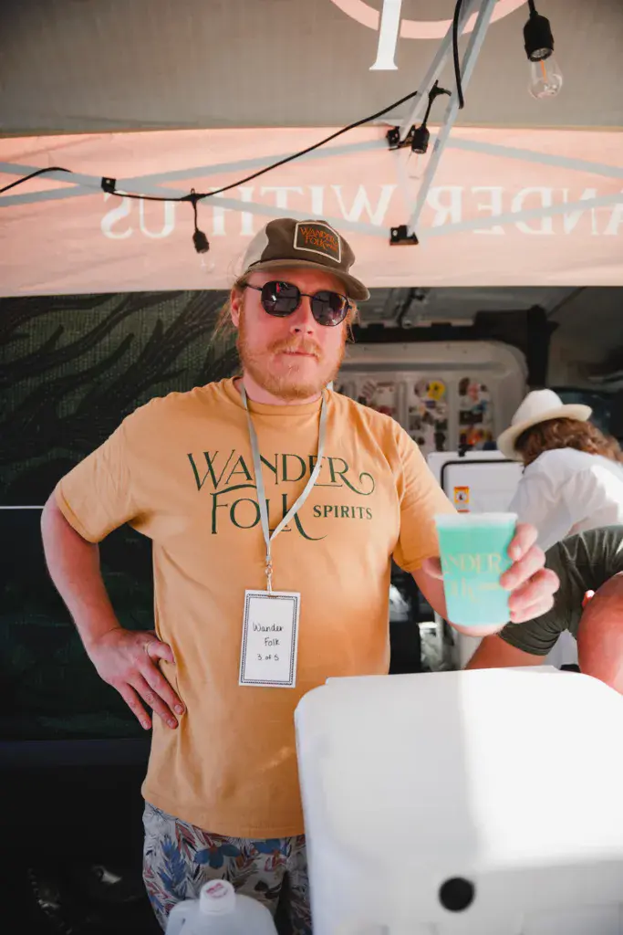 A festival staff member wearing a Wandro Folk Spirits branded shirt and lanyard holds a blue drink at an outdoor event.