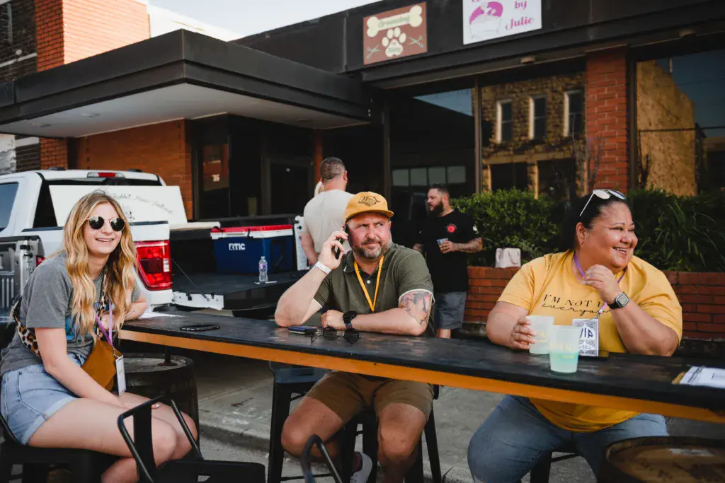 Three people sit at an outdoor table at a festival venue with brick buildings in the background.