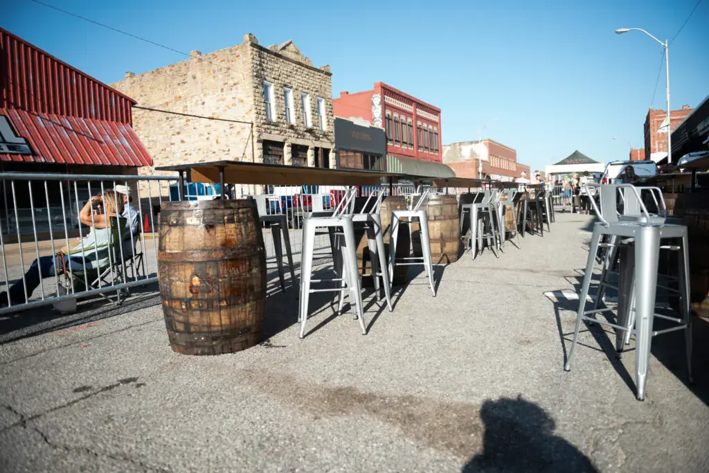Outdoor festival seating area with bar-height tables and stools arranged in front of historic downtown brick buildings under clear blue sky.