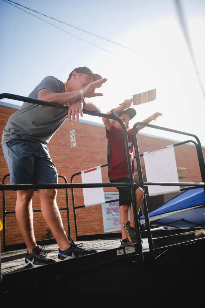 Two people stand on a stage platform gesturing expressively against a brick building backdrop.