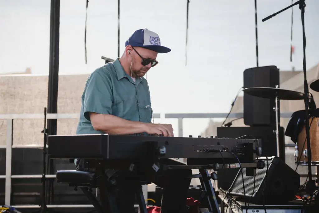 A performer plays keyboard on stage at an outdoor music festival under a cloudy sky.