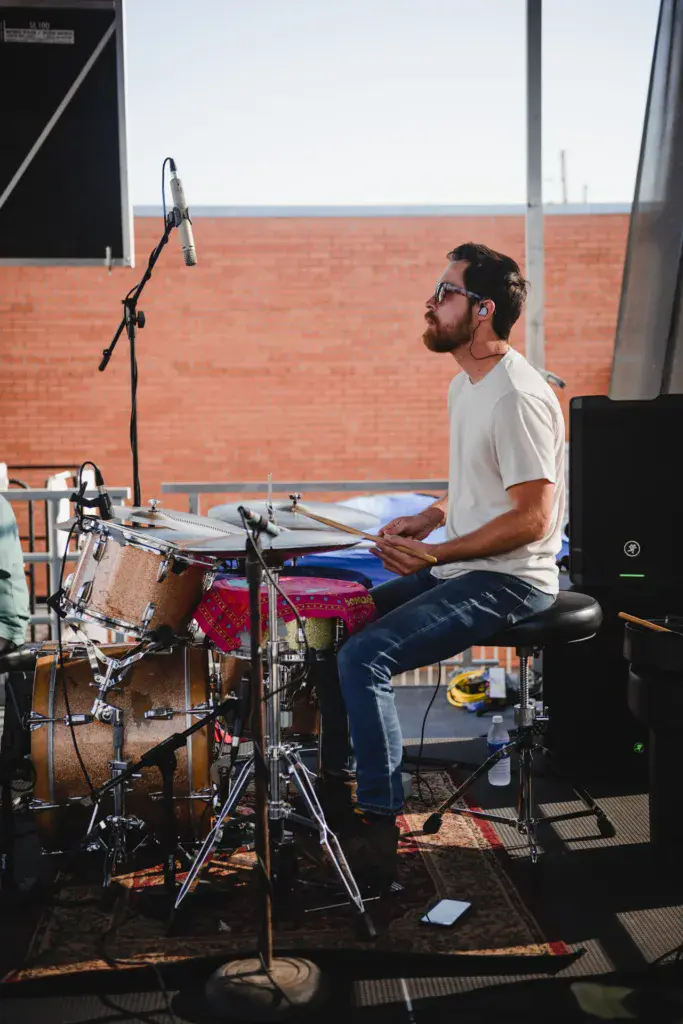 A drummer performing on stage at an outdoor music festival with a brick building backdrop.