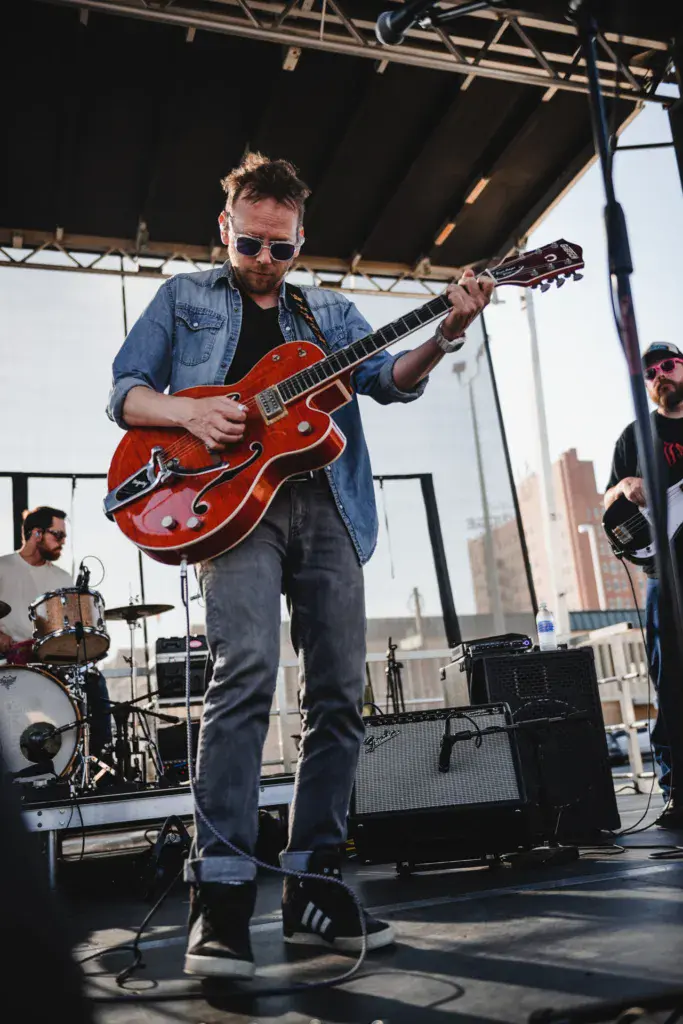 Guitarist with red semi-hollow body electric guitar performing on outdoor stage under metal canopy structure.