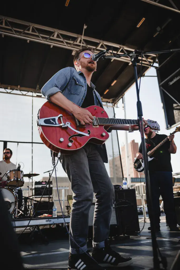 Musician wearing sunglasses and denim shirt plays a red semi-hollow electric guitar on an outdoor stage at night.
