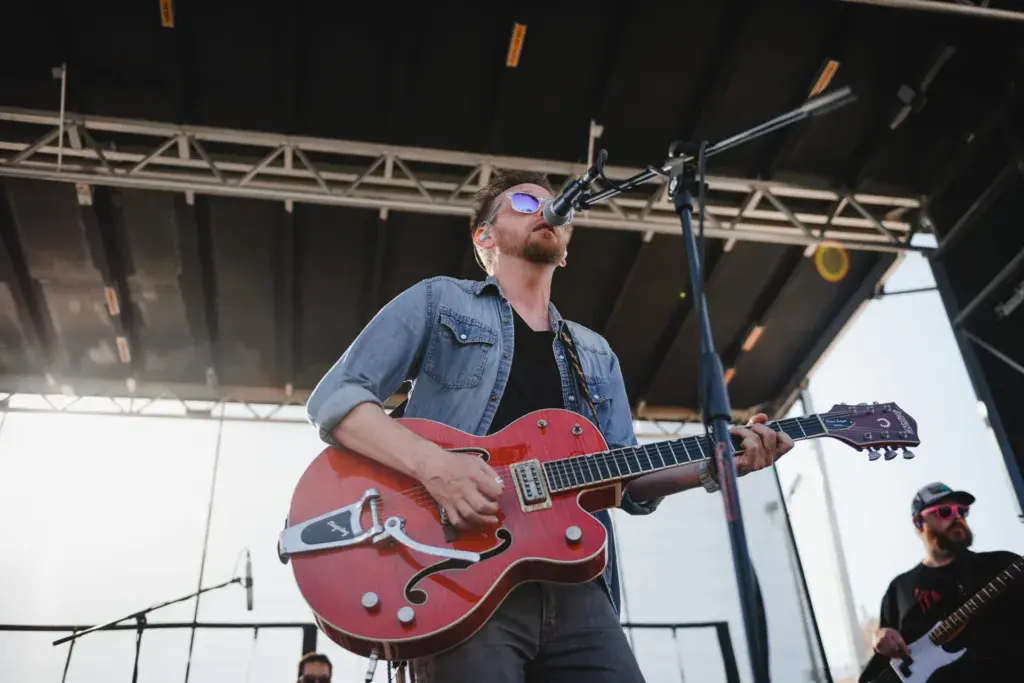 A guitarist performs on stage at the Dancing Rabbit Music Festival, wearing sunglasses and a denim shirt while playing a red semi-hollow body guitar.