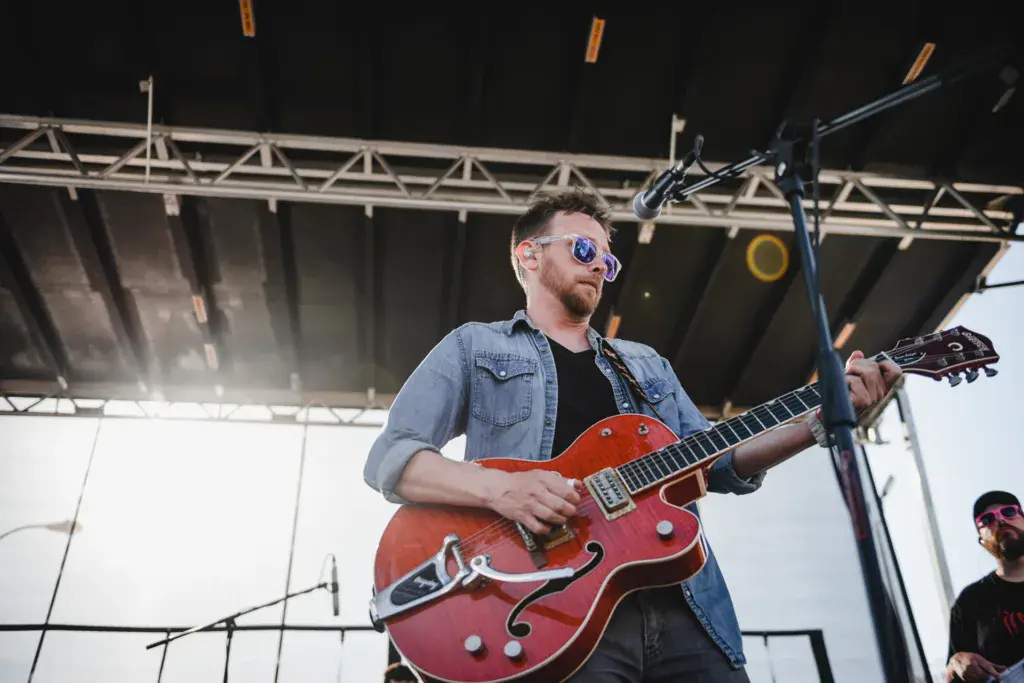 A guitarist wearing sunglasses and a denim shirt performs with a red semi-hollow body electric guitar on an outdoor festival stage.