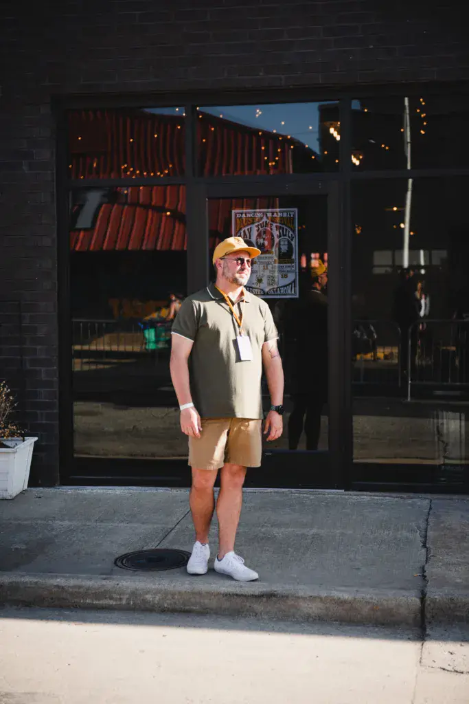 A person in a yellow cap and tan shorts stands outside a brick venue entrance with red curtains and event signage visible through the doors.