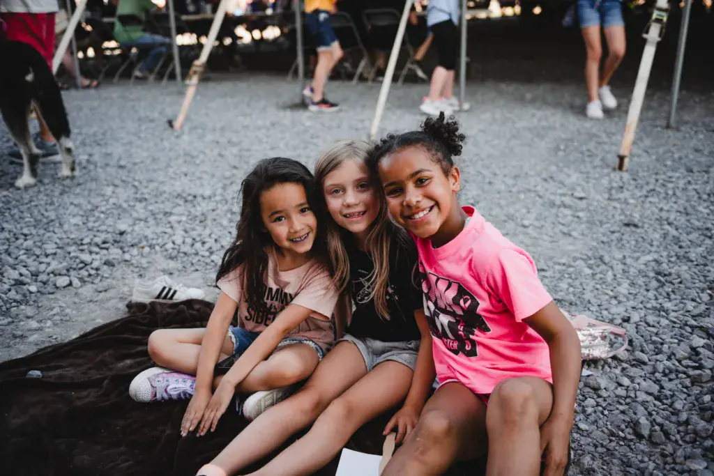 Three young children smile while sitting together on the ground at an outdoor festival venue.