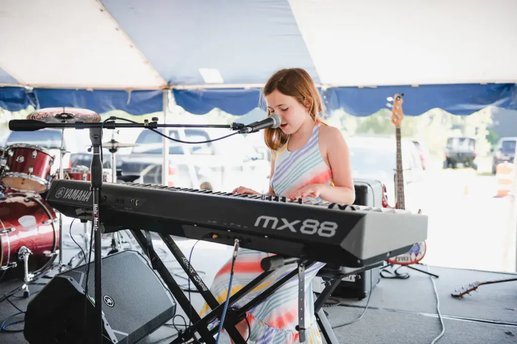 A performer plays keyboard and sings into a microphone on an outdoor festival stage under a blue and white tent.
