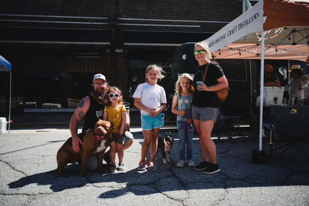 A group of adults and children with two dogs pose together at an outdoor festival vendor area.