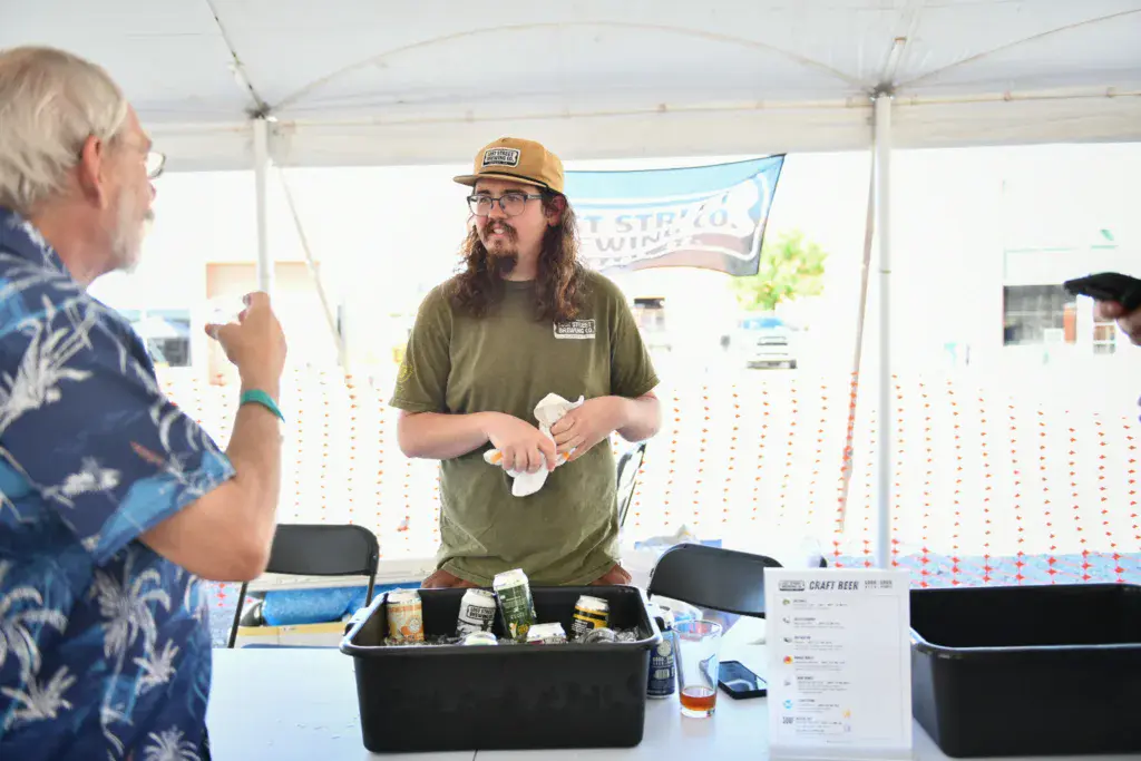 A vendor in a tan cap and olive shirt serves food from a booth under a white tent at an outdoor festival.