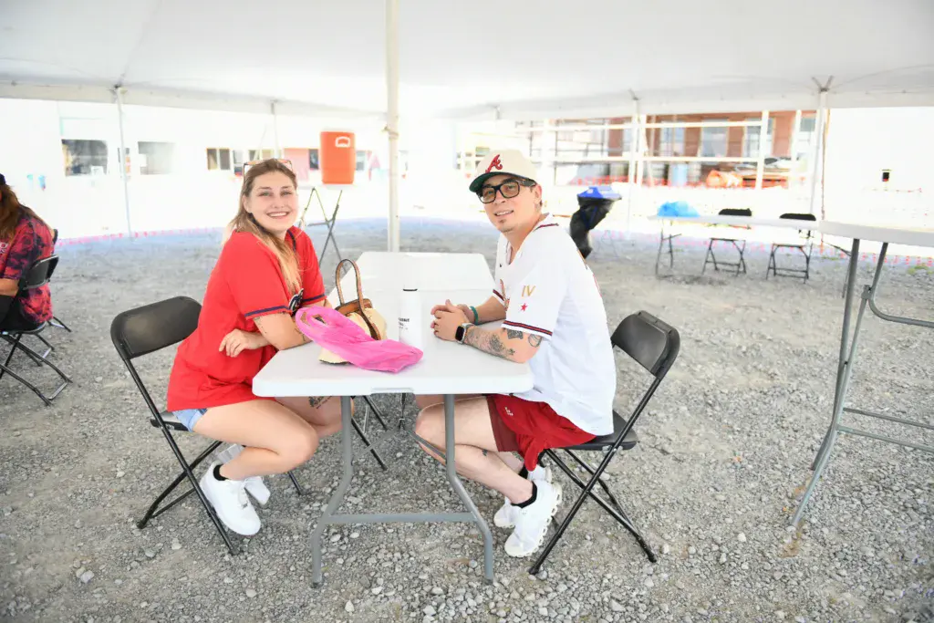 Two people smiling at a white table under a tent at an outdoor festival venue.