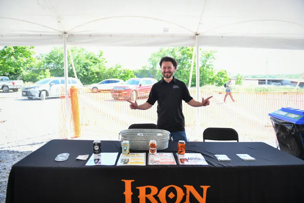 A person stands behind a sponsor booth table under a white tent at an outdoor festival venue.