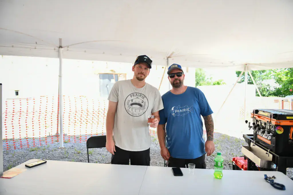 Two people wearing Prairie-branded shirts stand at a table under a white tent at an outdoor festival.