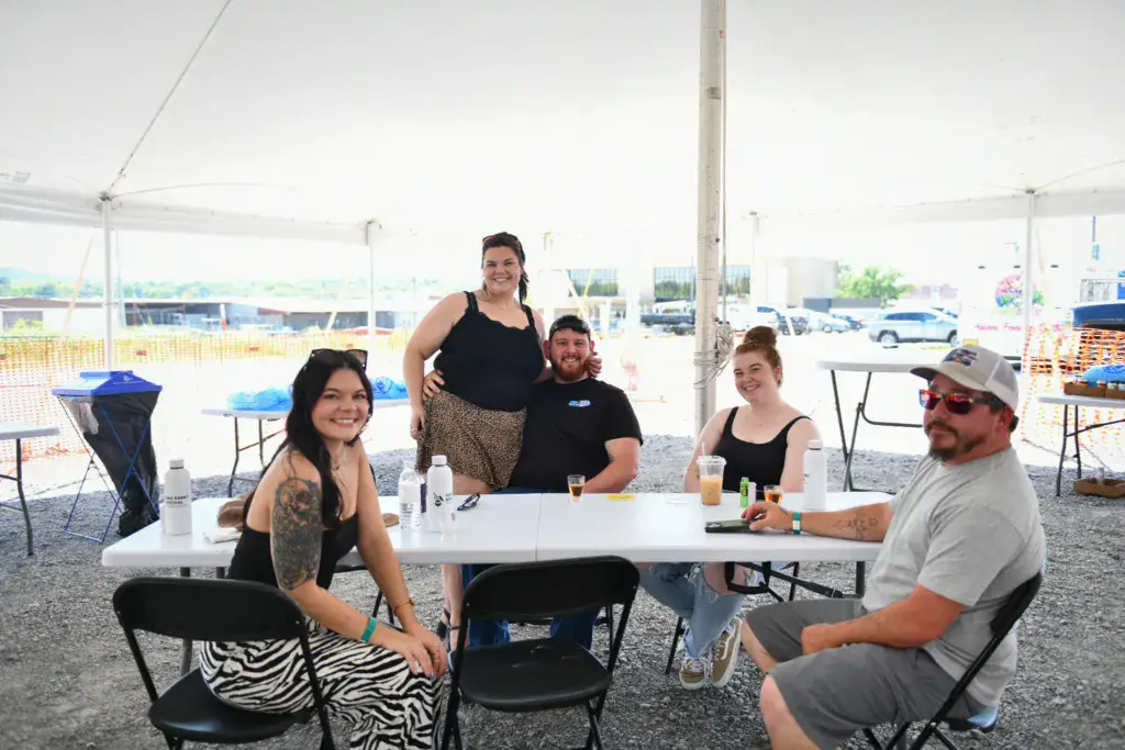A group of six people sitting and standing around a table under a white tent at an outdoor festival venue.