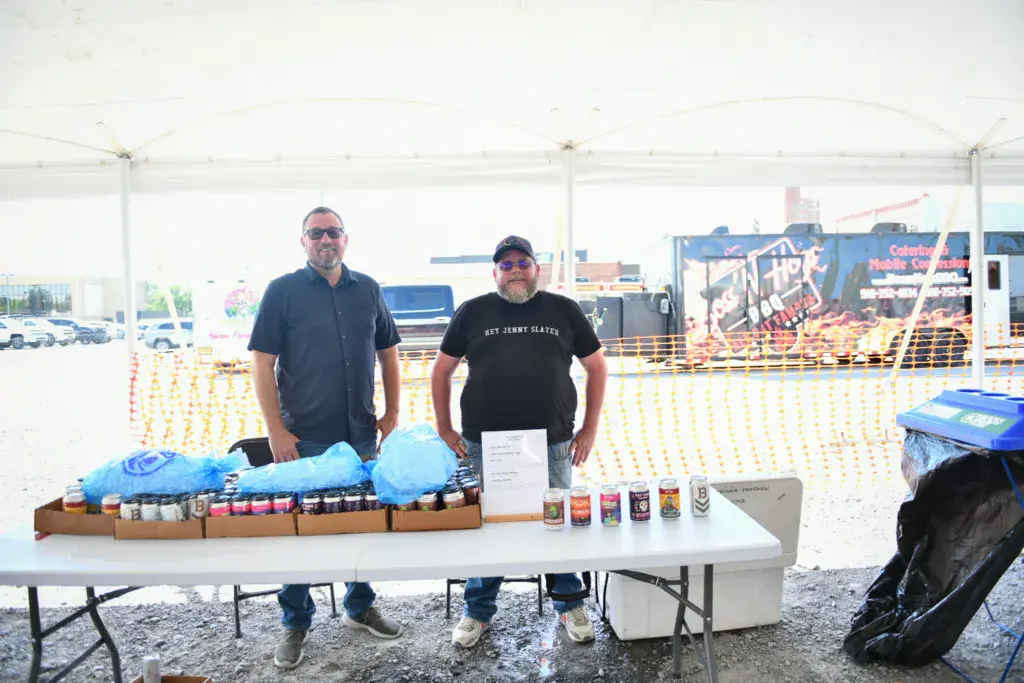 Two men stand behind a vendor table displaying merchandise and food items under a tent at an outdoor festival.