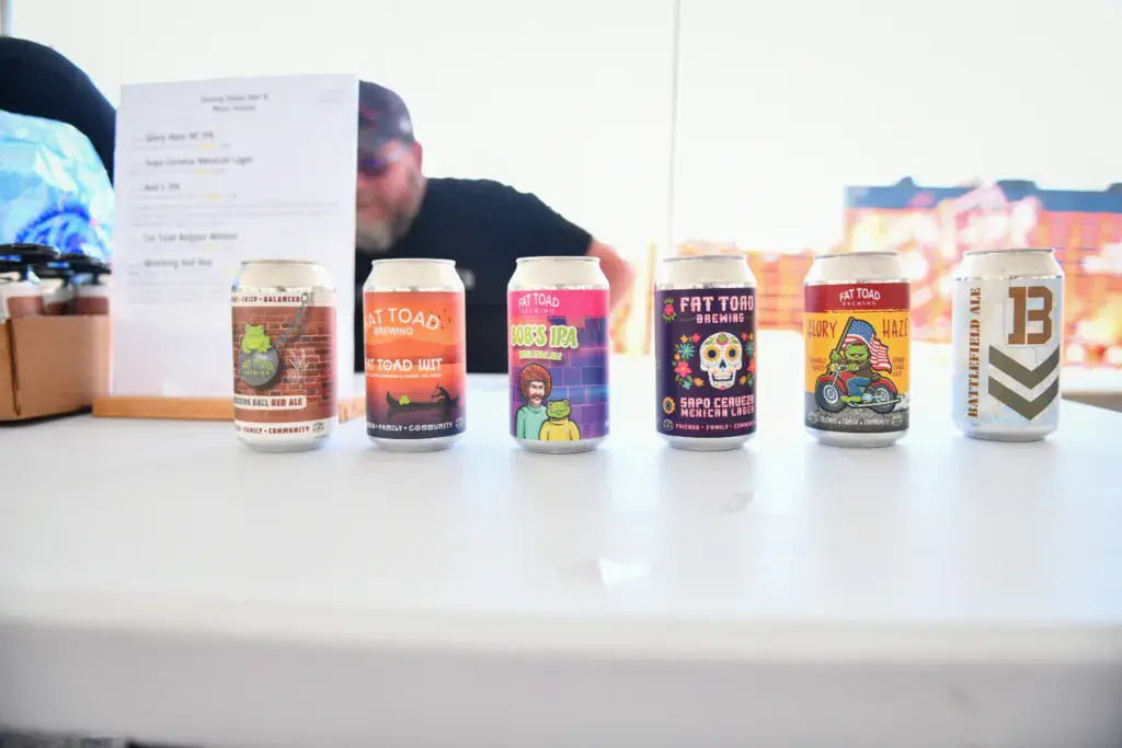 A row of colorful beer cans displayed on a white table with a person and signage visible behind them.