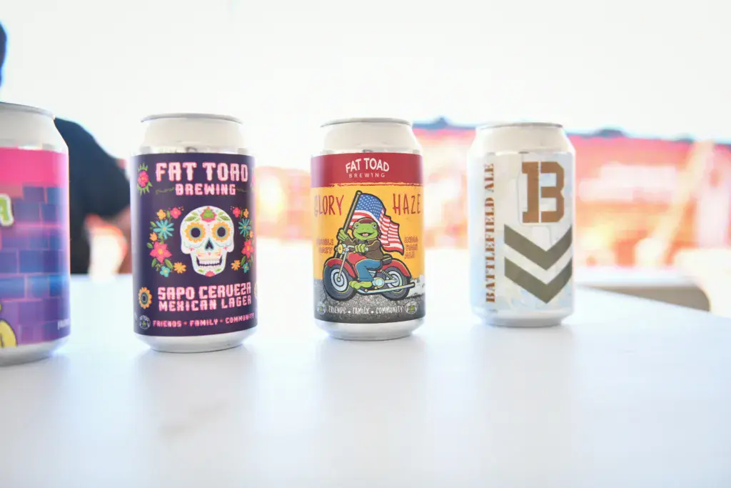 Several colorful beer cans from Fat Toad Brewing displayed on a white surface with a blurred outdoor background.