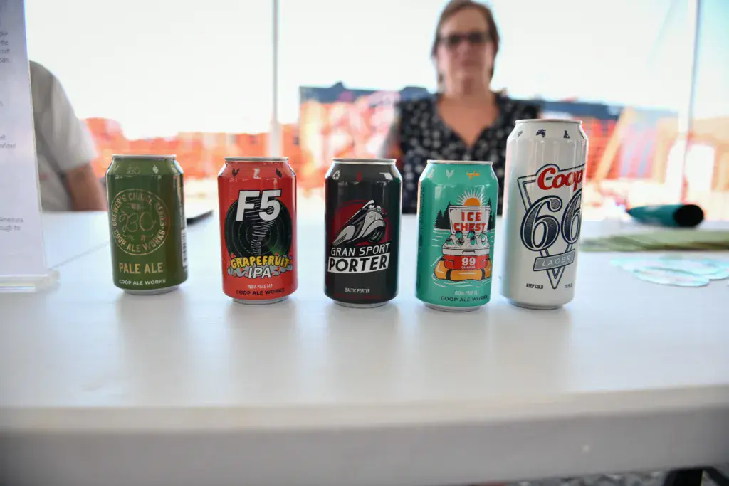 Five beer cans displayed on a white table at an outdoor festival with a person and colorful banners in the background.