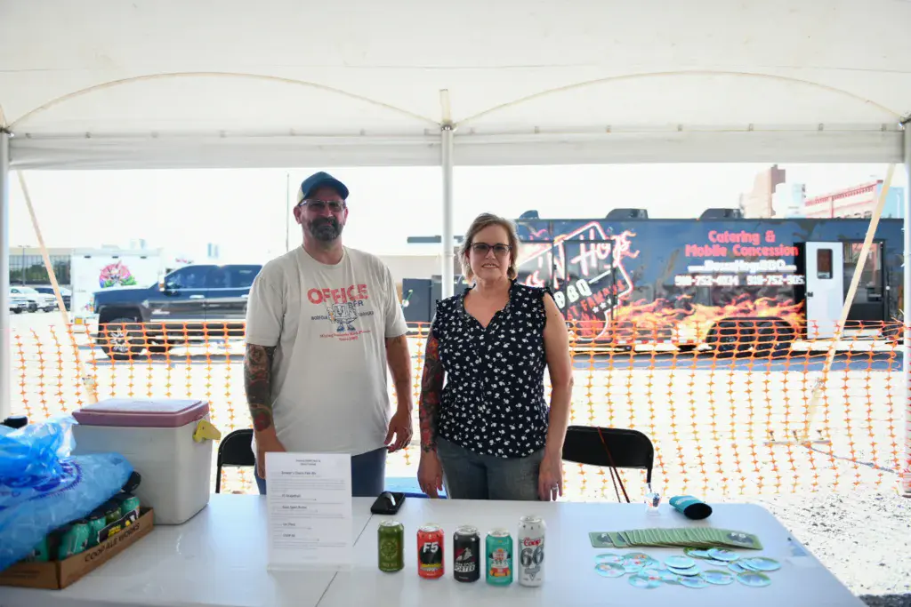 Two people standing behind a table with beverages at an outdoor festival venue under a white tent.