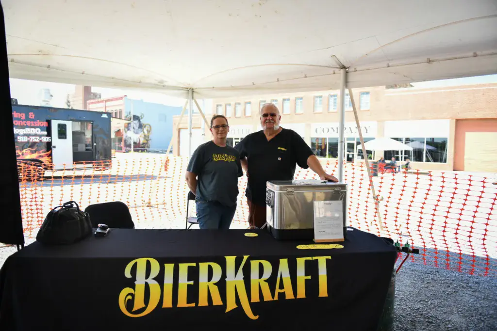Two people stand behind a Bierkraft branded table under a tent at an outdoor festival venue.