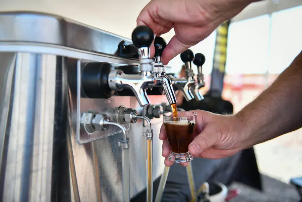A hand holds a glass being filled with dark beer from a tap on a stainless steel keg at an outdoor festival.