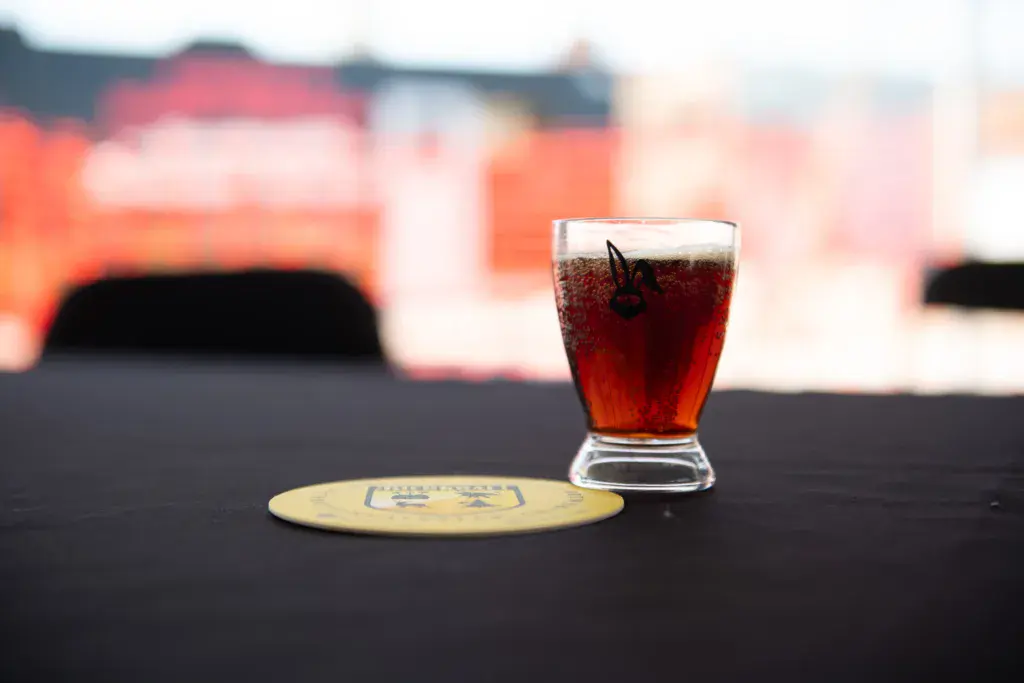 A glass of dark cola beverage sits on a dark surface with a coaster, stage structures blurred in the background.