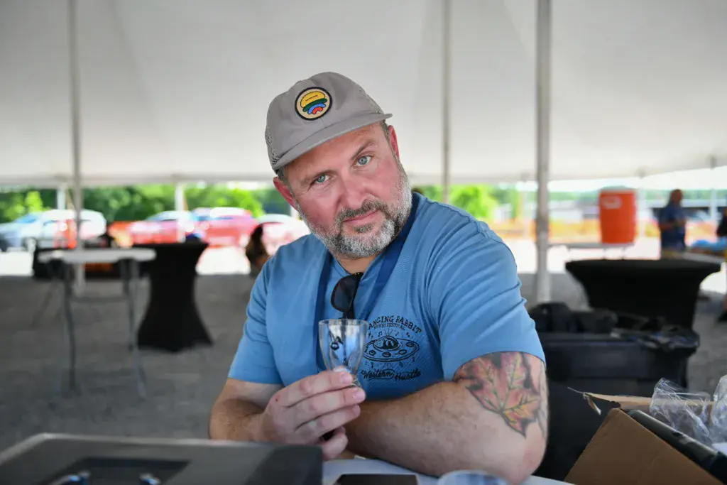 A man wearing a gray cap and blue shirt holds a drink while sitting under a festival tent.