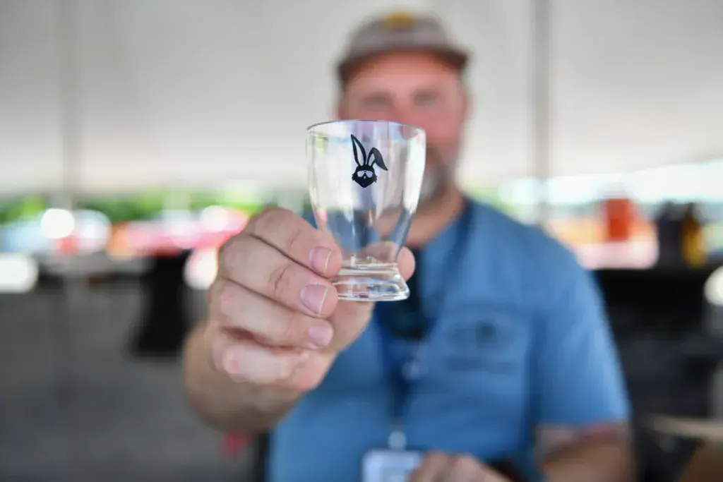 A person in a blue shirt and cap holds up a clear glass with a rabbit logo at an outdoor festival.