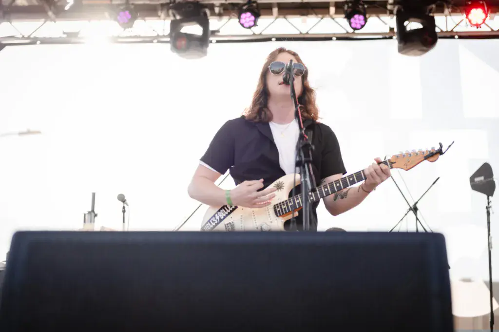 Performer wearing sunglasses playing an electric guitar at a music festival stage with stage lights overhead.
