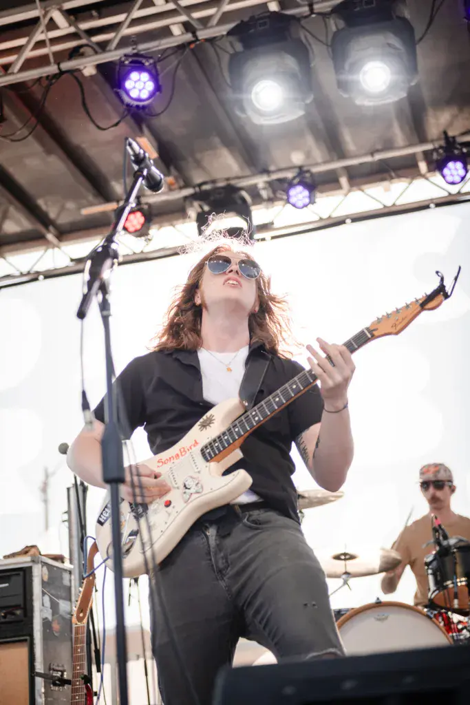 A guitarist wearing sunglasses performs on stage with a microphone, stage lights and drums visible behind them.