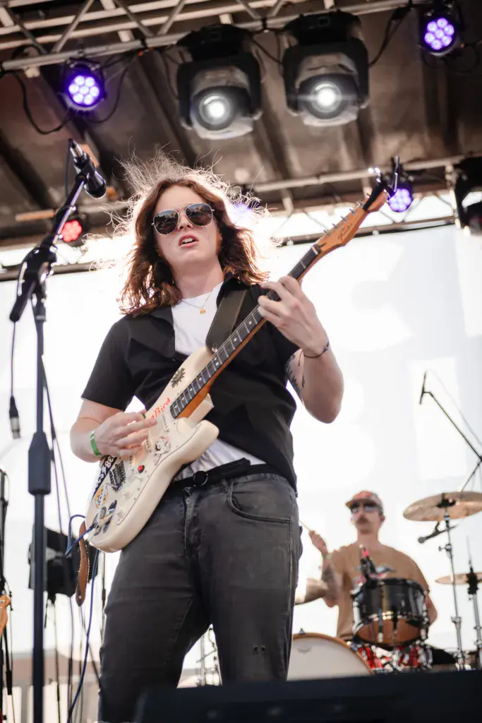 Guitarist performing on stage with sunglasses, raising one arm while playing a cream-colored electric guitar under purple and white stage lights.