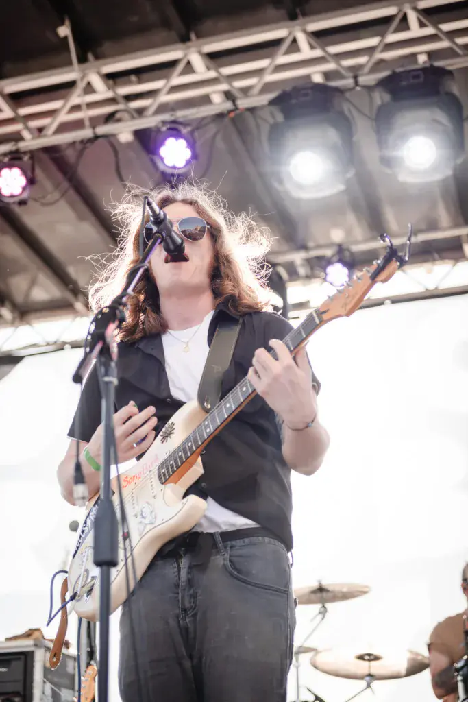 A musician wearing sunglasses performs on stage with an electric guitar under bright stage lights.