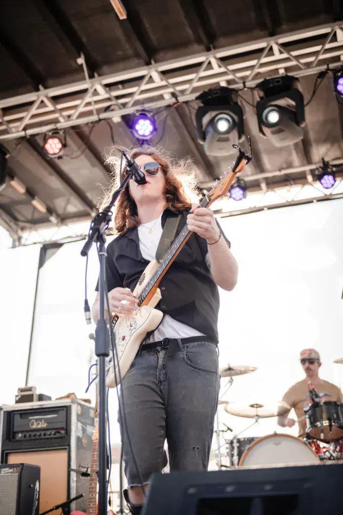 A musician with reddish hair wearing sunglasses plays guitar and sings into a microphone on stage under professional lighting.