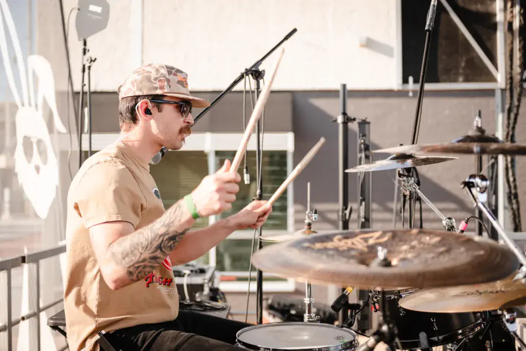 A drummer performs at an outdoor stage, playing a full drum kit in bright daylight.