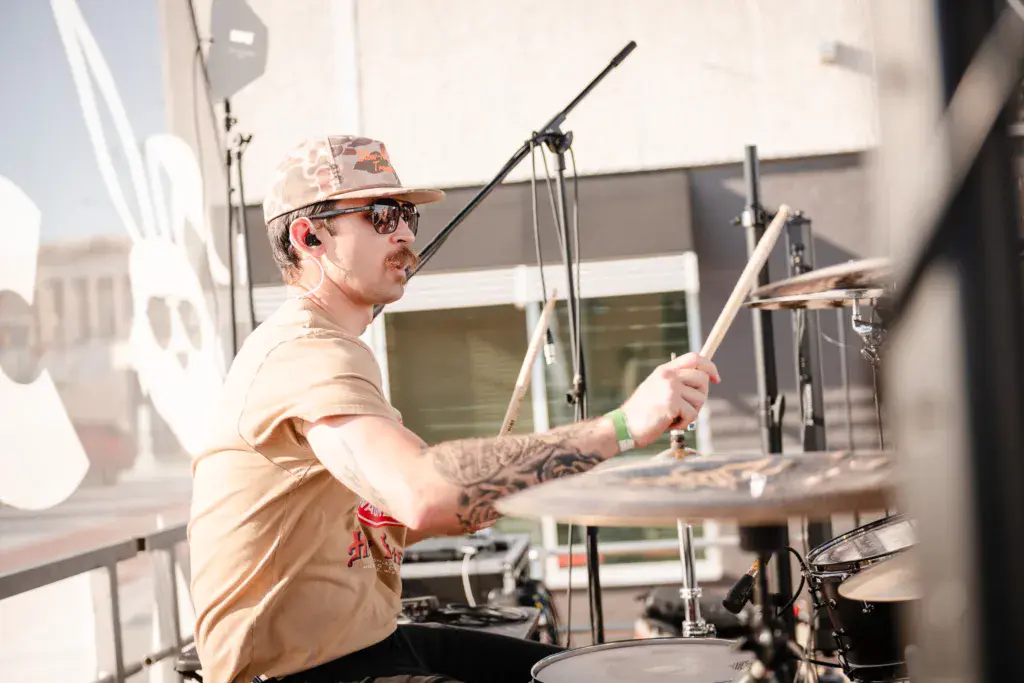 A drummer performs on stage wearing a tan shirt, cap, and sunglasses while playing a drum kit.