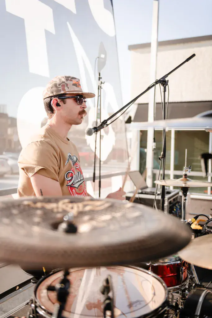 A drummer performs at an outdoor venue, seated behind drum kit with microphone and bright sunlight casting shadows.