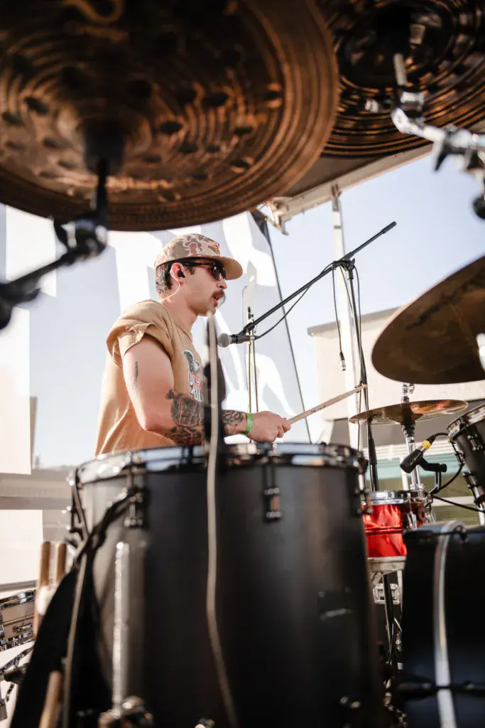 A drummer wearing a tan cap and sunglasses performs at an outdoor stage with cymbals and drum kit visible.