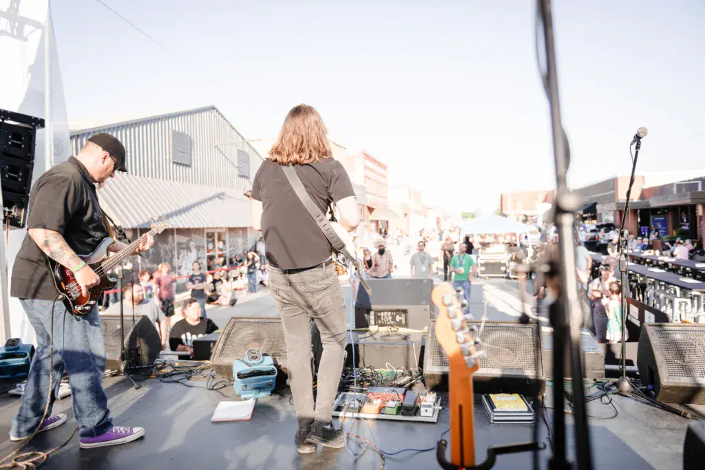 Two musicians perform on an outdoor stage with a large crowd gathered in the festival venue below.