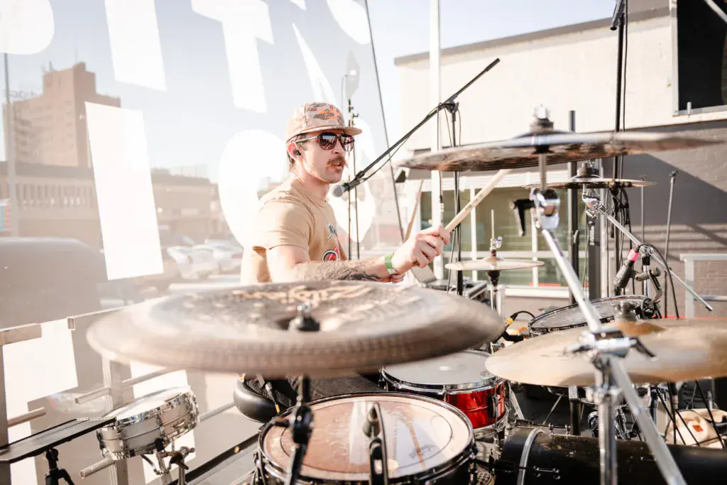 Drummer performs on stage at an outdoor music festival during daytime.