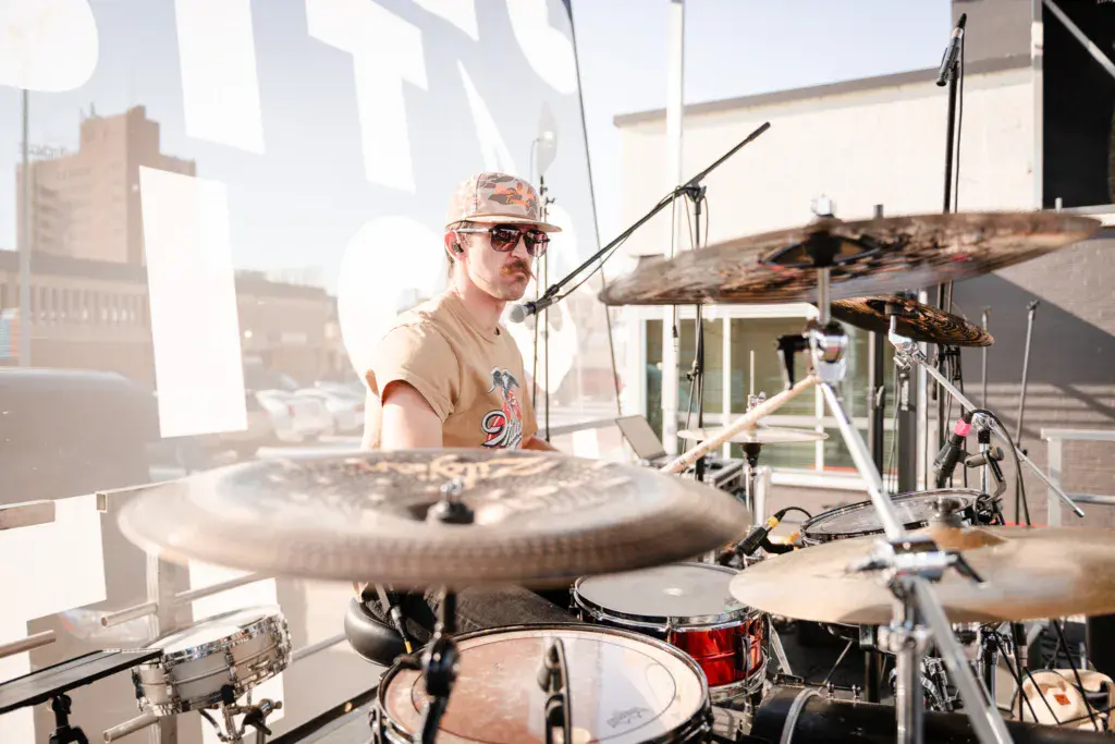 Drummer performing on stage at an outdoor music festival, wearing sunglasses and a tan cap.